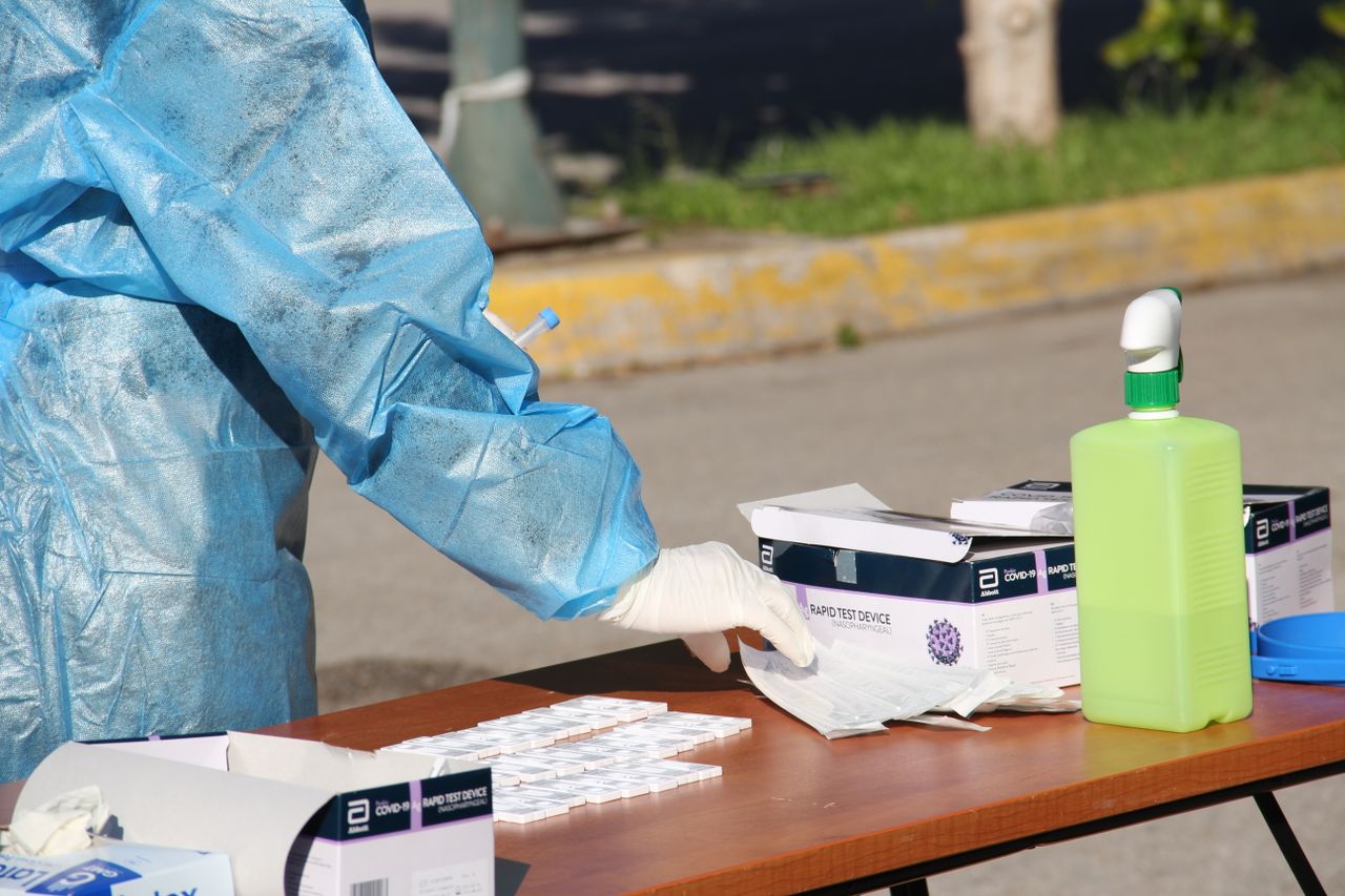 hero-img-02 Healthcare worker in protective gear conducting rapid COVID-19 testing outdoors. PPE and test kits visible.